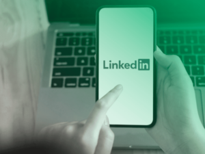 Pessoa acessando o aplicativo do LinkedIn em um smartphone, com um notebook ao fundo.