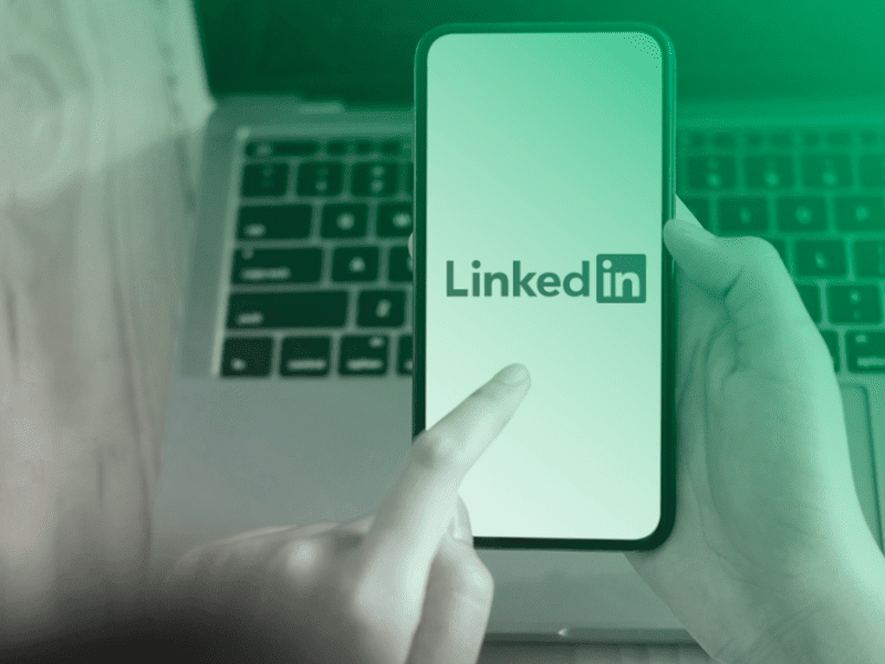 Pessoa acessando o aplicativo do LinkedIn em um smartphone, com um notebook ao fundo.